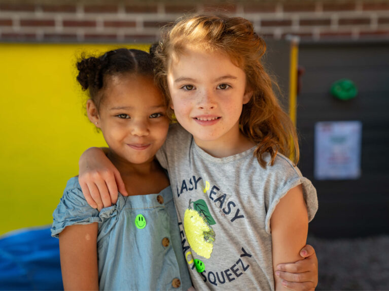 Duas meninas em idade pré-escolar com os braços sobre os ombros uma da outra