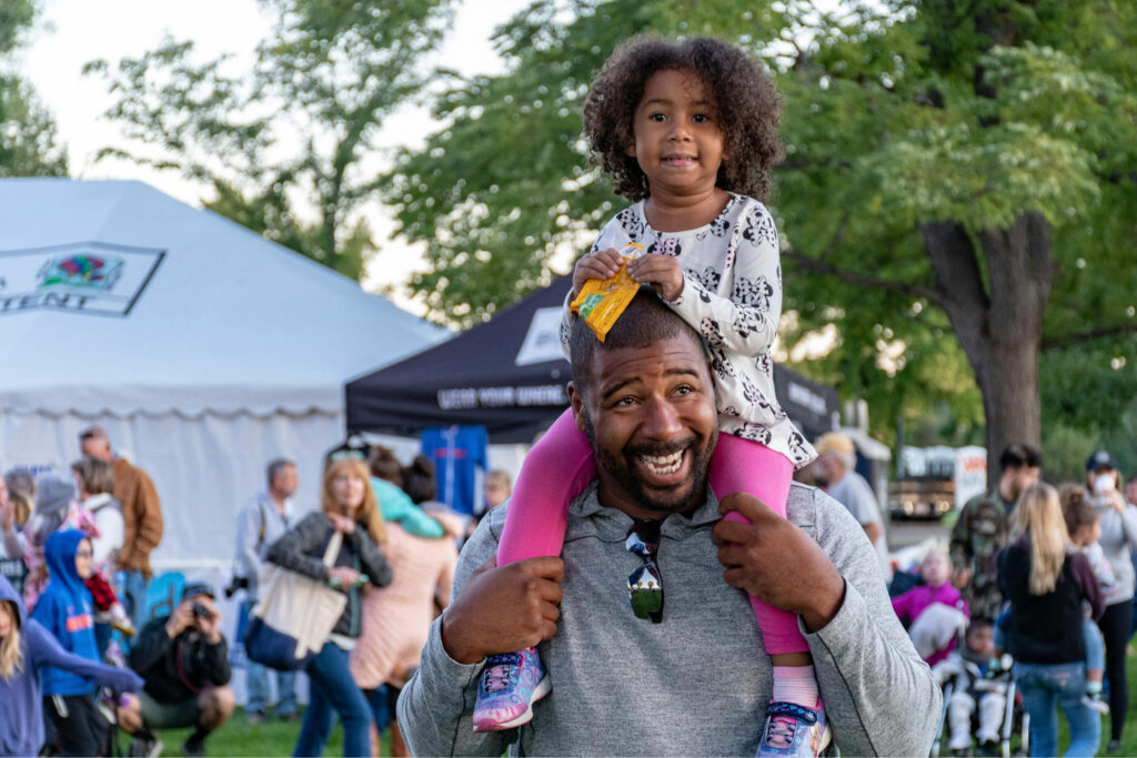 Pai afro-americano com a filha nos ombros em um festival comunitário.