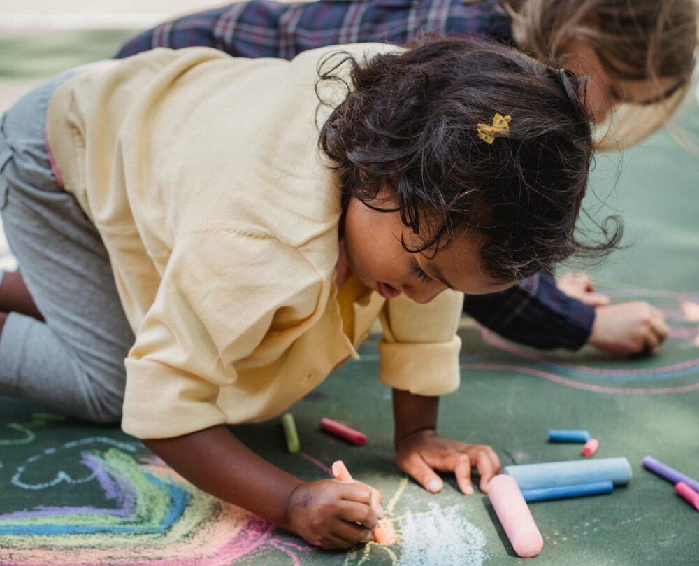 Young girl in yellow shirt drawing on cement with sidewalk chalk.