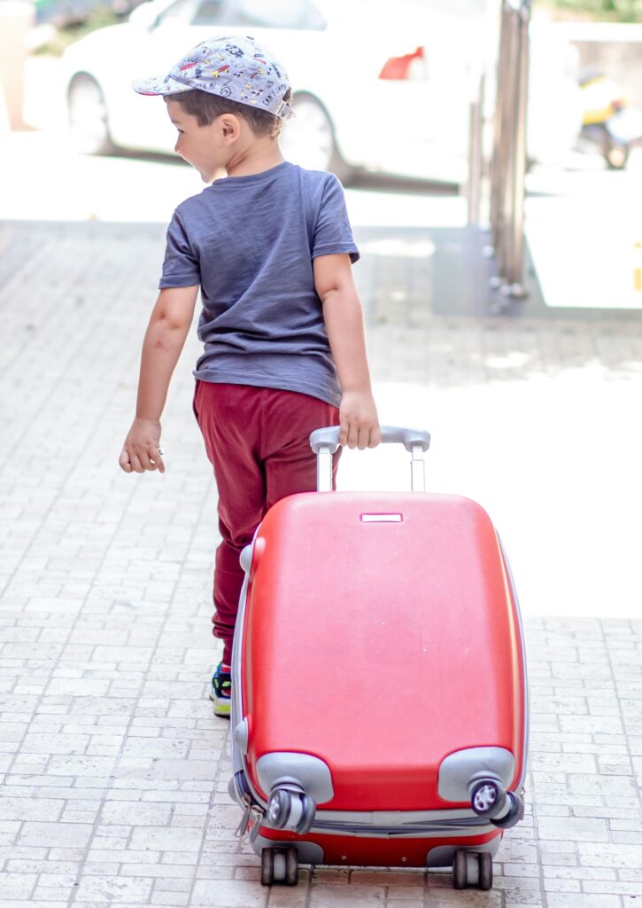 Niño con sombrero de pelota caminando y tirando de una maleta roja.