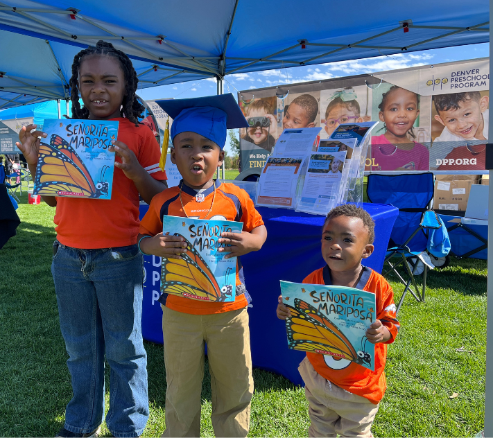 Three children holding books in front of a tent