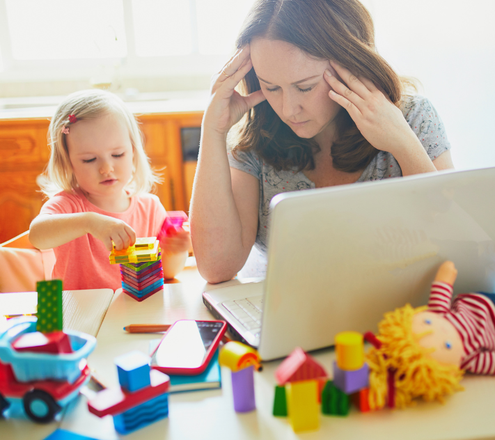 Mother holding her head while sitting in front of computer; young girl playing with magna tiles.