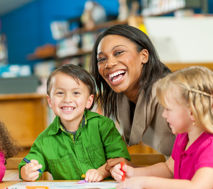 Young boy and female teacher smiling while young girl draws.