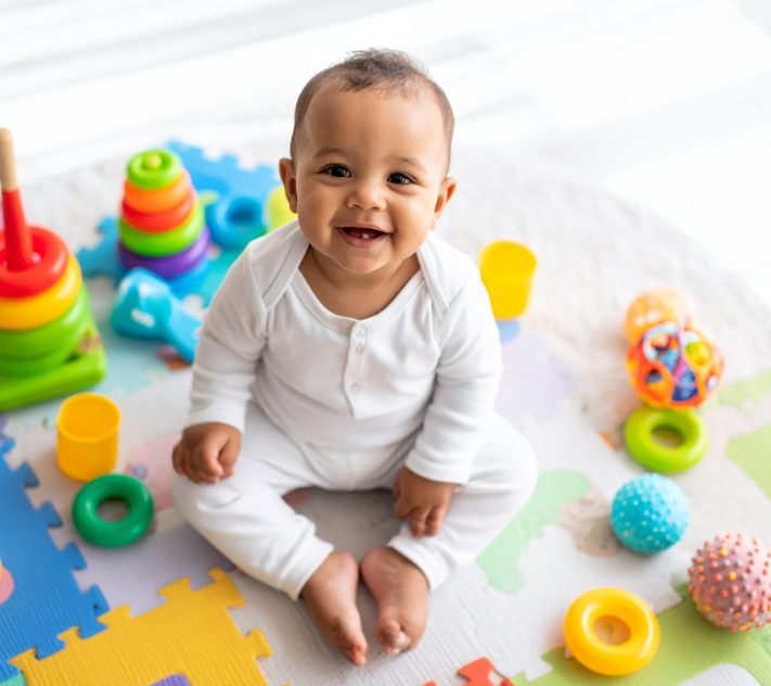 Bebé sonriendo mientras está sentado en el suelo rodeado de juguetes.
