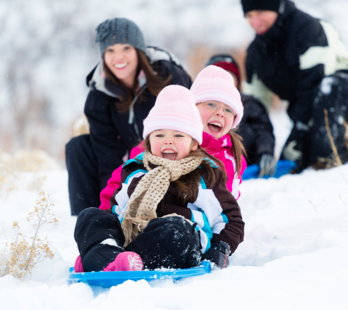Deux jeunes filles descendent une colline en luge, leur mère en arrière-plan.