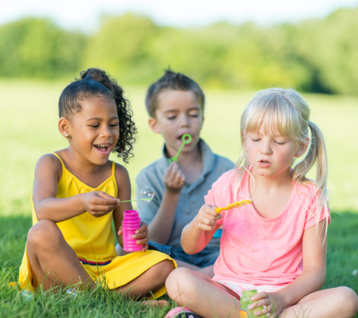 Three young kids sitting in the grass while blowing bubbles.