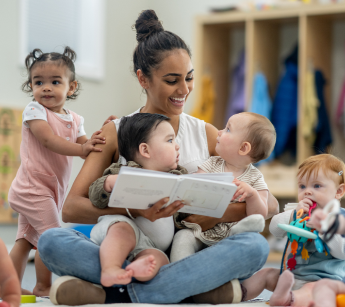 Une enseignante assise par terre, tenant un tout-petit et un livre, entourée d'autres tout-petits.